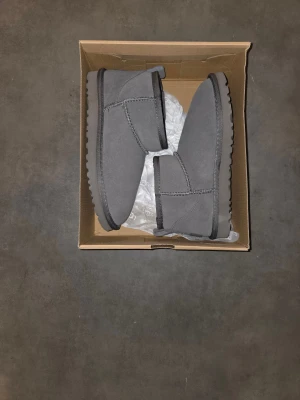 Grå UGG boots i mocka - Mysiga grå boots från UGG i mjuk mocka med foder av päls. Skorna har en låg modell med rund tå och platt sula, perfekt för kyliga dagar. Klassisk UGG-logga bak på hälen och dragflik för enkel påtagning.