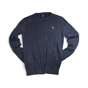 Mörkblå kabelstickad tröja från GANT - Snygg mörkblå kabelstickad tröja från GANT i premium bomull. Tröjan har rund hals, ribbade muddar och broderad logga på bröstet. Perfekt för dig som gillar klassisk stil med en modern touch.