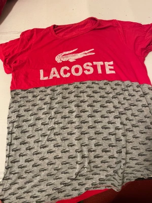 Röd och grå Lacoste t-shirt - Säljer en snygg t-shirt från Lacoste med röd överdel och grå nederdel. Framsidan har stort Lacoste-tryck och krokodillogga, medan den grå delen är täckt av små svarta krokodiler. Klassisk passform och korta ärmar. Perfekt för dig som gillar streetstyle och loggor.