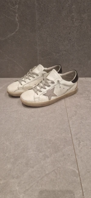 Golden Goose vita sneakers med stjärna - Golden Goose sneakers i vitt skinn med grå mockadetaljer och en ikonisk stjärna på sidan. Skorna har grå snörning, rund tå och svart hälparti. Snyggt slitna med vintagekänsla och platt sula. Perfekta för dig som gillar streetstyle och unika detaljer.( rätt säker på att de är fejk.) De är hål vid tån och har ett jacka på insidan av skon.