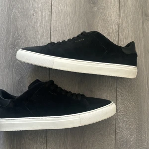 Svarta sneakers från Axel Arigato - Snygga svarta sneakers från Axel Arigato med stilren design. Skorna har en rund tå, platt vit sula och är tillverkade i mocka med detaljer i skinn. Klassisk snörning och minimalistisk look som passar till jeans eller dressade byxor.