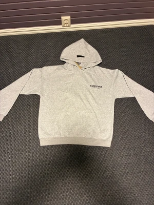 Grå Essentials Fear of God hoodie - Snygg grå hoodie från Essentials Fear of God med svart logotyp på bröstet. Tröjan har huva, ribbade muddar och en klassisk känguruficka framtill. Perfekt för dig som gillar streetwear och vill ha en clean look. PASSAR DIG SOM Ä CA 175-185