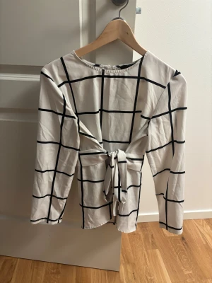 Vit rutig blus med knyt från SHEIN - Säljer en vit blus från SHEIN med svart rutigt mönster. Blusen har långa ärmar och snygg knytning framtill som ger en cool touch. Materialet är mjukt och lätt polyester, perfekt för dig som gillar stilrena och trendiga plagg.
