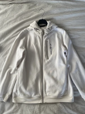 Vit hoodiejacka från Sail Racing - Vit hoodiejacka från Sail Racing med dragkedja framtill och bröstficka med logga. Jackan har justerbar huva med snörning och långa ärmar. Perfekt för dig som gillar stilren och sportig look.