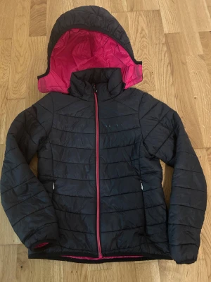 Svart pufferjacka från Everest - Svart pufferjacka från Everest med rosa foder och dragkedja. Jackan har huva, två sidofickor med dragkedja och quiltad design. Perfekt för kyliga dagar och har en snygg kontrast mellan det mörka yttertyget och den rosa insidan.
