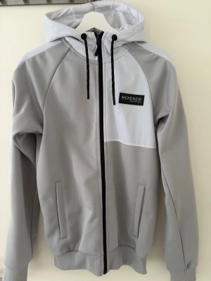 Ljusgrå vindjacka från McKenzie - Snygg ljusgrå vindjacka från McKenzie med huva och svarta snören. Jackan har hel dragkedja framtill, två fickor och en stilren logga på bröstet. Tillverkad i polyester och perfekt för dig som gillar en clean look.