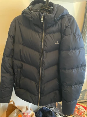 Mörkblå pufferjacka från GANT - Säljer en mörkblå pufferjacka från GANT med huva och dragkedja. Jackan har ett stilrent snitt, diskret logga på bröstet och coolt tryck på insidan. Perfekt för kalla dagar och riktigt snygg till streetwear-stilen.