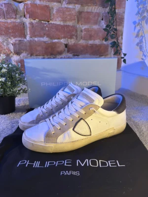 Vita Philippe Model sneakers - Snygga vita sneakers från Philippe Model med gråa detaljer och mörkblå häl. Skorna har klassisk snörning, rund tå och är tillverkade i skinn med mockadetaljer. Perfekta för dig som gillar stilrena och trendiga sneakers med exklusiv känsla.