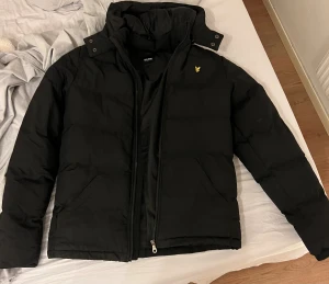 Svart pufferjacka från Lyle & Scott - Svart pufferjacka från Lyle & Scott med gul logga på bröstet. Jackan har hög krage, dragkedja och två sidofickor. Perfekt för kalla dagar med sin tjocka vaddering och stilrena look.