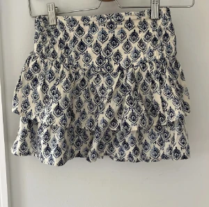 Vit och blå mönstrad kjol från Zara - Säljer en supersöt kort kjol från Zara i vitt med blått mönster. Kjolen har volanger och resår i midjan. Köpt i en Zara butik i Portugal.