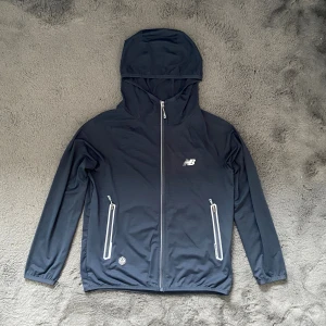 Mörkblå vindjacka från New Balance - Snygg mörkblå vindjacka från New Balance med huva och hel dragkedja framtill. Jackan har två fickor med dragkedja och vita detaljer, samt elastiska muddar vid ärmslut. Perfekt för blåsiga dagar och sportiga outfits. PRISET KAN DISKUTERAS 