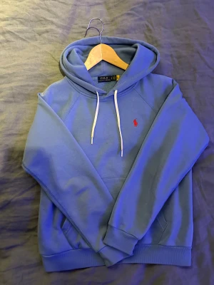 Blå hoodie från Polo Ralph Lauren - Snygg blå hoodie från Polo Ralph Lauren med klassisk röd broderad logga på bröstet. Tröjan har huva med vita snören och ribbade muddar vid ärmslut och nederkant. Perfekt för chill dagar och enkel att matcha med jeans eller joggers. Passar 160