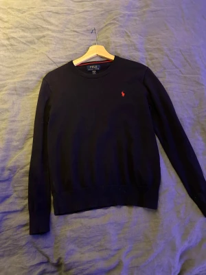 Mörkblå tröja från Polo Ralph Lauren - Säljer en mörkblå tröja från Polo Ralph Lauren med klassisk rund hals och röd broderad logga på bröstet. Tröjan är långärmad och har ribbade muddar vid ärmslut och nederkant. Perfekt för dig som gillar stilrena och tidlösa plagg.