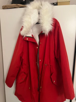 Röd parkasjacka med fuskpäls - Röd parkasjacka med vit fuskpäls runt huvan och insidan. Jackan har dragkedja, knappar, snörning i midjan och stora fickor framtill. Perfekt för kalla dagar när du vill sticka ut lite extra.