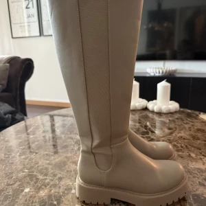 Beige höga boots med platt sula - Snygga beige boots med högt skaft och chunky platt sula. Skorna har rund tå och är tillverkade i ett slätt syntetmaterial med elastiska paneler på sidorna för extra komfort. Perfekta för att lyfta din outfit med en trendig touch.