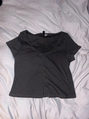 Mörkgrå ribbad topp med knappar - En mörkgrå ribbad topp med korta ärmar och djup rundringning. Toppen har en rad små knappar framtill som ger en snygg detalj. Perfekt att styla med jeans eller kjol för en chill look. Helt ny och oanvänd men tagit bort prislappen!