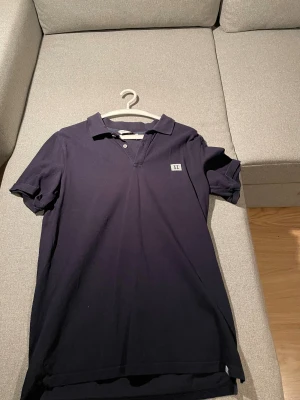 Mörkblå piké från Les Deux - Snygg mörkblå pikétröja från Les Deux med klassisk krage och två knappar framtill. På bröstet finns en broderad patch med romerska siffran II. Materialet är mjuk bomull och passformen är normal. Perfekt för dig som gillar stilrena och enkla plagg.