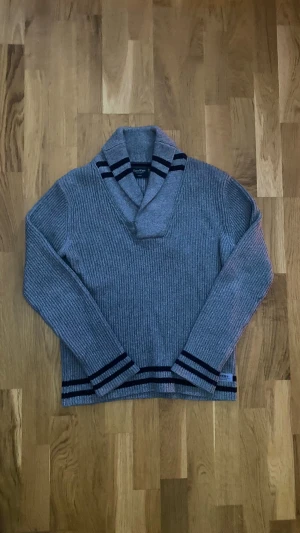 Grå stickad Cardigan från East West - Snygg grå kofta från East West i ribbstickad design med svarta ränder på krage, ärmslut och nederkant. Koftan har bred krage med två knappar och långärmad passform. Perfekt för dig som gillar klassisk stil med en twist. Storlek M och passar mig som är 175cm perfekt! 