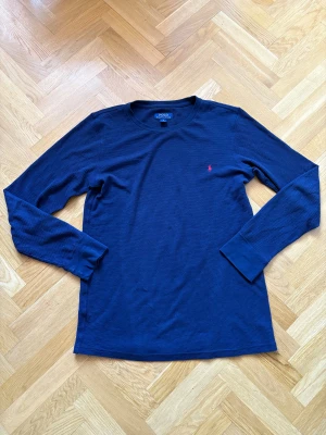 Marinblå långärmad tröja Polo Ralph Lauren - Snygg marinblå långärmad tröja från Polo Ralph Lauren i storlek L. Tröjan är tillverkad i bomull med ett ribbat, stickat tyg och har den klassiska röda Polo-loggan broderad på bröstet. Perfekt för dig som gillar stilrena och tidlösa plagg.