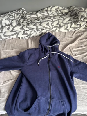 Marinblå hoodie från Polo Ralph Lauren - Säljer en marinblå hoodie från Polo Ralph Lauren med dragkedja och vit snörning i huvan. Tröjan har långa ärmar, fickor framtill och den klassiska röda Polo-loggan broderad på bröstet. Perfekt för chill dagar eller när du vill vara extra comfy.