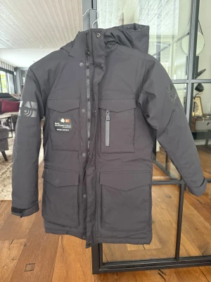 Svart vinterparkas Sail Racing - Svart parkas från Sail Racing med huva, flera stora fickor och snygga logodetaljer på bröstet och ärmen. Jackan är vadderad och har dragkedja samt knappar framtill. Perfekt för kalla dagar när du vill hålla stilen och värmen.