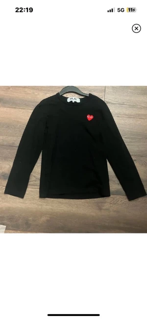 Svart långärmad tröja Comme des Garçons - Svart långärmad tröja från Comme des Garçons PLAY med ikoniskt rött hjärta på bröstet. Tröjan har en clean och enkel design med rund hals och normal passform. Perfekt för dig som gillar stilrena plagg med en twist.