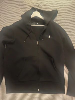 Svart hoodie från Polo Ralph Lauren - Svart hoodie från Polo Ralph Lauren med klassisk vit logga på bröstet. Tröjan har huva med snörning, dragkedja framtill och två stora fickor. Perfekt för chill dagar och enkel att matcha med jeans eller joggers.