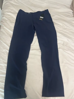 Mörkblå kostymbyxor Jack Nicklaus - Snygga mörkblå kostymbyxor från Jack Nicklaus med rak passform och klassisk design. Byxorna har en gul broderad björn vid fickan och är gjorda i polyester och elastan för skön stretch. Perfekta för dig som gillar stilrena och bekväma byxor.