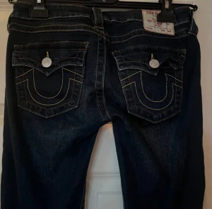 True Religion mörkblå bootcut jeans - Säljer ett par mörkblå jeans från True Religion med bootcut passform. Jeansen har snygga kontrastsömmar, fickor med lock och silverfärgade knappar. Modellen heter Joey och har låg midja. Storlek W27