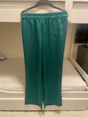 Gröna satinbyxor från & Other Stories - Snygga gröna byxor i glansigt satinmaterial från & Other Stories. Modellen har hög midja med resår, raka och vida ben samt praktiska fickor i sidorna. Perfekt för dig som gillar en chill och trendig look.