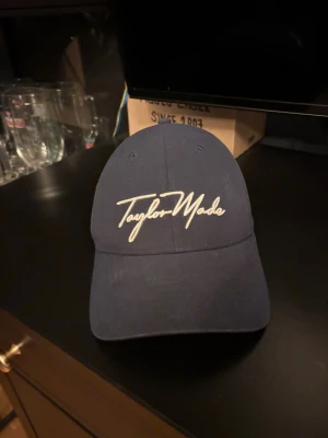 Mörkblå TaylorMade keps - Snygg mörkblå keps från TaylorMade med broderad logga framtill och bak. Klassisk böjd skärm och justerbar rem baktill för perfekt passform. Perfekt för golf eller chill vardagsstil. Materialet känns som bomull och kepsen har ventilerande hål på ovansidan.
