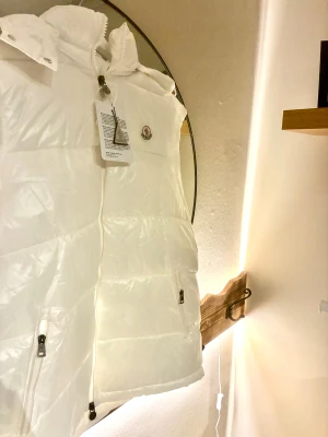 Vit dunväst  - Snygg vit dunväst i storlek 5XL (xl/xxl) Västen har en hög krage, huva och två fickor med dragkedja. Quiltad design och klassisk logga på bröstet. Perfekt för att addera en clean vibe till din outfit.