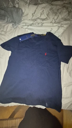 Mörkblå t-shirt från Polo Ralph Lauren - Snygg mörkblå t-shirt från Polo Ralph Lauren i mjuk bomull. Klassisk rund hals och korta ärmar. På bröstet finns den ikoniska röda Polo-loggan som ger en clean och stilren look. Perfekt för dig som gillar enkel och tidlös design.