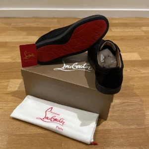Christian Louboutin svarta sneakers mocka - Svarta sneakers från Christian Louboutin i lyxig mocka med ikonisk röd sula. Klassisk låg modell med rund tå och snörning. Kommer med originalkartong och dustbag. Perfekta för dig som vill sticka ut med exklusiv stil.