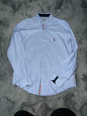 Ljusblå skjorta från Polo Ralph Lauren - Passar även S klassisk krage och broderad logga på bröstet. Skjortan har långa ärmar, vita knappar och coola rödblå detaljer vid kragen och nederkant. Tillverkad i mjuk bomull för skön känsla och stilren look.
