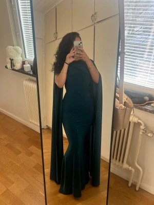 Mörkgrön långklänning med cape - Säljer en mörkgrön långklänning med cape-detalj och figurnära passform. Klänningen har en asymmetrisk axel och svepande tyg som ger en dramatisk look. Materialet är mjukt och stretchigt, vilket gör att den sitter snyggt och bekvämt. Perfekt för bal eller fest! Klänningen har en pytte liten defekt pga en nål som legat där men ingen som syns i helhet (se sista bild). Annars är klänningen i mycket fint skick! Priset går att diskhos snabb affär 