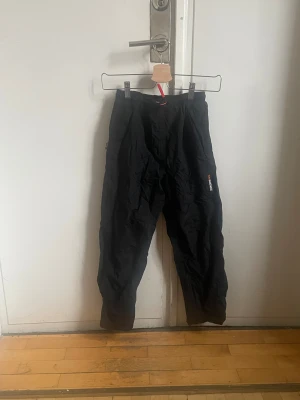 Svarta täckbyxor från Didriksons - Svarta täckbyxor från Didriksons 1913 med broderad logga på benet och orange snörning i midjan. Byxorna har en loose passform och är gjorda i syntetmaterial som står emot väta och kyla. Perfekta för kalla dagar och vinteraktiviteter.