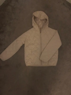 Beige quiltad tjej jacka med huva från verani - Quiltad beige jacka med huva och dragkedja framtill från verani. Jackan har ett stilrent rutmönster och är lätt vadderad, perfekt för kyliga dagar. Långärmad och i syntetmaterial som ger en lätt känsla. Enkel och clean look som passar till det mesta.