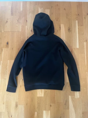 Svart hoodie från Lyle & Scott - Svart hoodie från Lyle & Scott med klassisk gul logga på bröstet. Tröjan har huva med snörning, stor magficka och långa ärmar. Mjuk och skön polyesterkvalitet, perfekt för chill eller häng. Enkel och stilren design som funkar till det mesta.