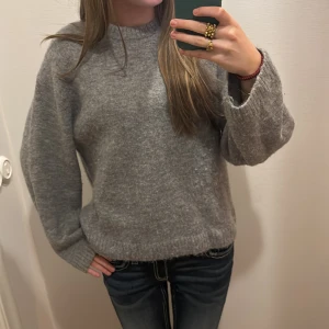 Grå stickad tröja med rund hals - Mysig grå stickad tröja med rund hals och långa ärmar. Tröjan har en relaxed fit och ribbade muddar vid ärmslut och nederkant. Perfekt för kyliga dagar och enkel att matcha med jeans eller kjol.