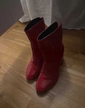 Röda boots med klack från Bershka - Snygga röda boots från Bershka i storlek 37. Skorna har en rund tå, blockklack och är tillverkade i glansigt skinnmaterial. De har dragkedja på insidan för enkel på- och avtagning. Perfekt för dig som vill sticka ut med färg och stil. Kedjan är lite trög men går ändå att använda som vanligt på ena skon annars är dom som nya knappt använda 