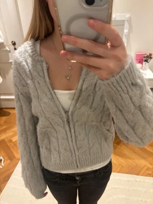 Ljusgrå stickad kofta från Zara - Mysig ljusgrå stickad kofta med dragkedja framtill och kabelstickat mönster. Koftan har långa ärmar och ribbade muddar vid ärmslut och nederkant. Står 152 i storlek men skulle säga ca 164 