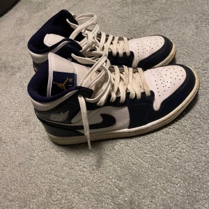 Nike Air Jordan 1 Mid blå och vita sneakers - Säljer ett par Nike Air Jordan 1 Mid sneakers i blått och vitt med klassisk design. Skorna har snörning, rund tå och platt sula. Perfekta för dig som gillar streetwear och vill sticka ut med en ikonisk modell.
