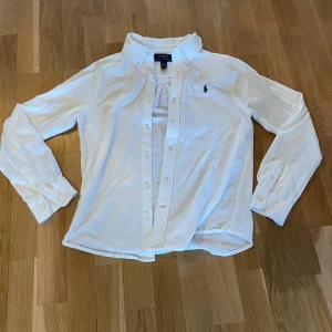 Vit skjorta från Polo Ralph Lauren - Klassisk vit skjorta från Polo Ralph Lauren med button down-krage och broderad logga på bröstet. Skjortan har långa ärmar, knappar framtill och är tillverkad i samma tyg som piké tröjor. Perfekt för en clean och stilren look.