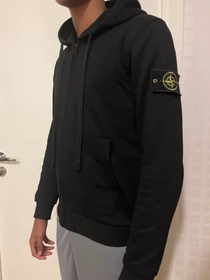Svart hoodie från Stone Island - Svart hoodie från Stone Island med klassisk patch på vänster ärm. Tröjan har huva med snörning, magficka och ribbade muddar. Perfekt för dig som gillar streetwear och vill ha en clean look med snygga detaljer.