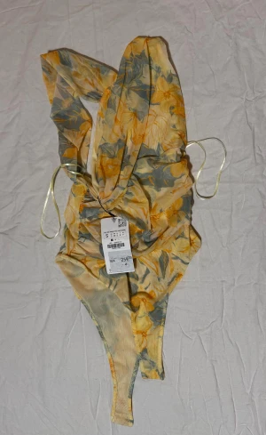 Blommig omlott body från Zara i ljusgult, aldrig använd strl S - Supersnygg body från Zara i ljusgult med blommigt mönster i gult och grått. Omlott-design framtill och öppen rygg ger en cool och trendig look. Materialet är mjukt och stretchigt syntet. Säljes med prislapp kvar.