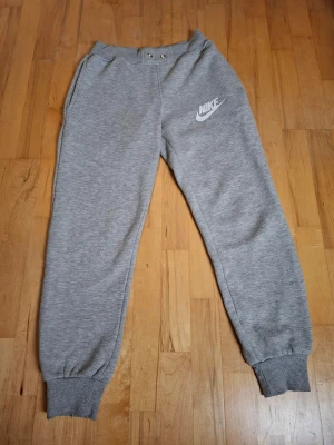 Grå mjukisbyxor från Nike, storlek S - Snygga grå mjukisbyxor från Nike med klassisk logga på vänster ben. Byxorna har ribbade muddar vid benslut, elastisk midja med snörning och två sidofickor. Perfekta för chill eller träning, tillverkade i mjukt bomullsmaterial för maximal komfort.