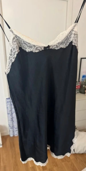 Svart spetsklänning Victoria's Secret XS - Svart kortklänning från Victoria's Secret med tunna axelband och vit spets längs urringning och nederkant. Söt liten rosett framtill och en liten slits med spetsdetalj. Klänningen har en slimmad passform och är perfekt för dig som gillar en feminin och chic stil.