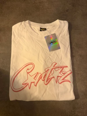 Vit Cartiz t-shirt med rosa tryck - Vit t-shirt från Cartiz med stort rosa tryck framtill i cool graffiti-stil. Klassisk rund hals och korta ärmar. Tillverkad i mjuk bomull, perfekt för en avslappnad streetwear-look. Snygg detalj med holografisk tag kvar. Passar M