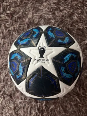 Champions League München 25 fotboll - Säljer en officiell Champions League München 25 finalfotboll från Adidas. Bollen har en snygg design med vita och mörkblå paneler, blå detaljer och stjärnmönster. Tillverkad i slitstarkt syntetmaterial, perfekt för matcher och träning.